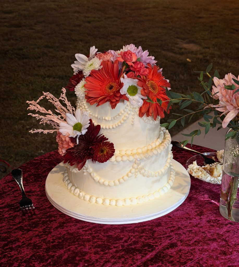 A two-tier white cake decorated with red gerbera daisies and piped scrollwork