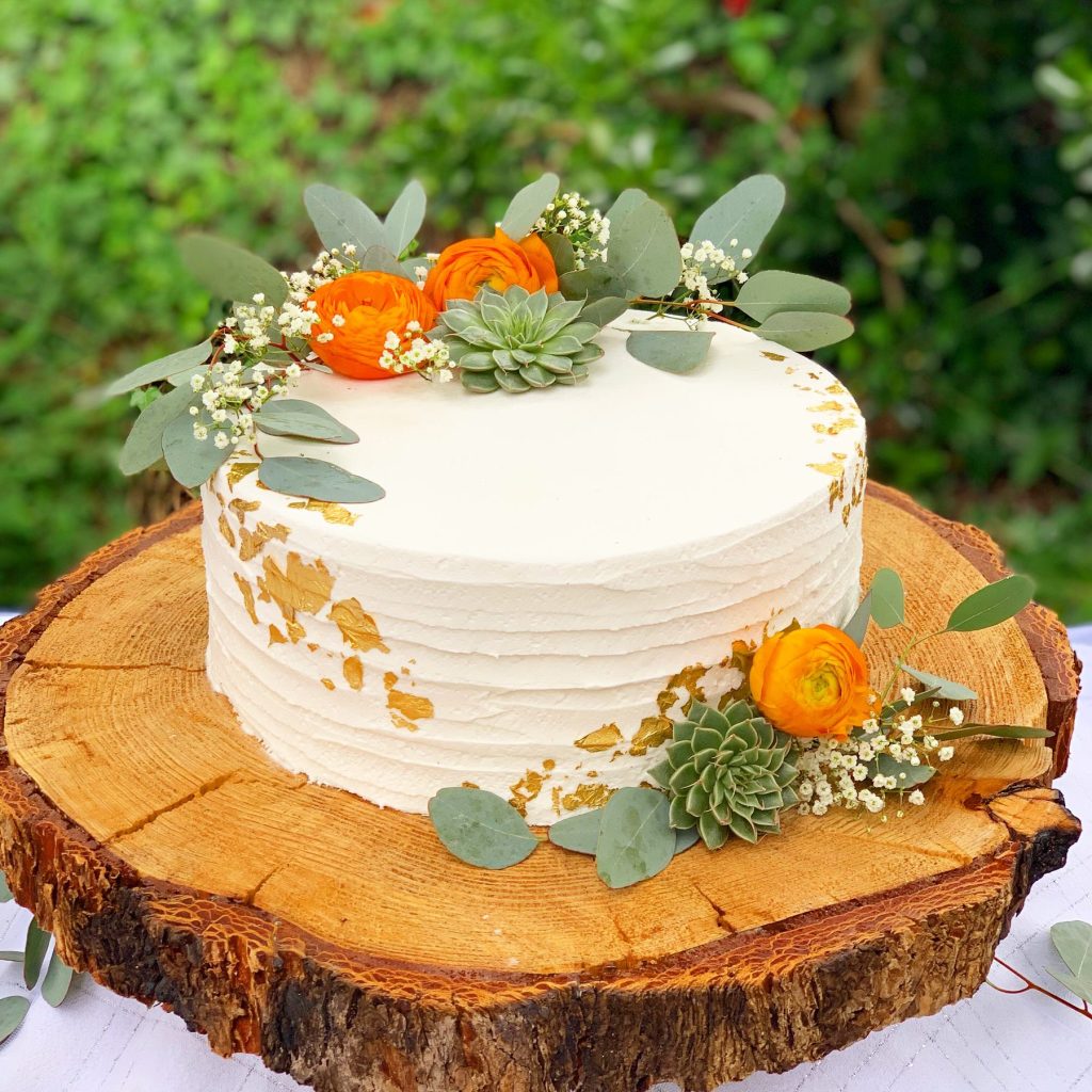 A white buttercream cake topped with orange ranunculus and eucalyptus on a wood slice