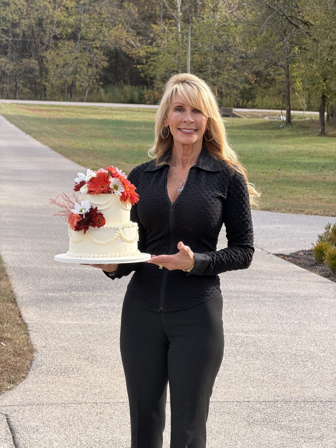 Timorie holding a two-tier white cake decorated with red and orange gerbera daisies